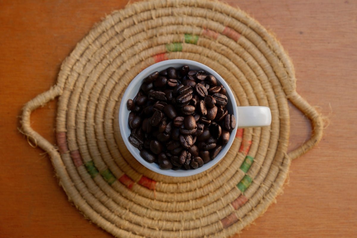 Coffee beans fill a white cup on a woven mat.