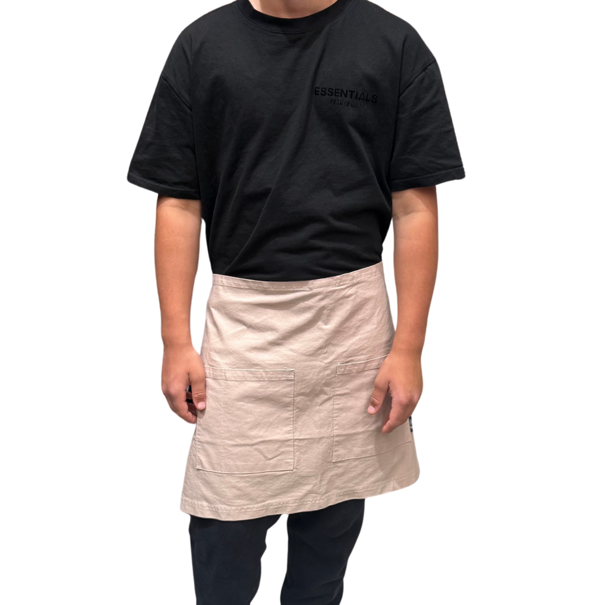 Person wearing a black t-shirt with 'essentials' text and a beige apron on a white background