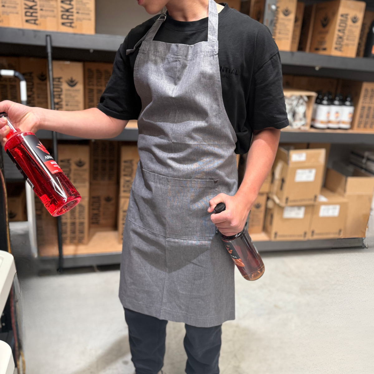 Person wearing a gray apron holding two bottles in a warehouse setting with cardboard boxes and shelves.