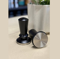Thumbnail for Two black coffee tampers on a light surface with a blurred plant in the background