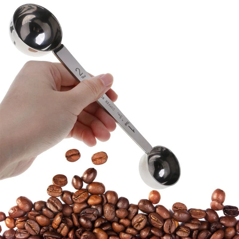 Hand holding a stainless steel measuring spoon over coffee beans on a white background