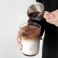 Thumbnail for A person pouring dark coffee from a stainless steel pitcher into a clear glass of iced milk coffee, with a white wall in the background.