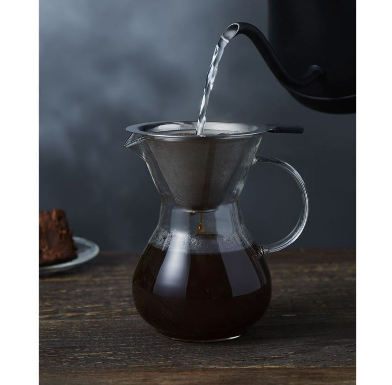 Pouring liquid into a glass carafe with a metal filter on a wooden surface.